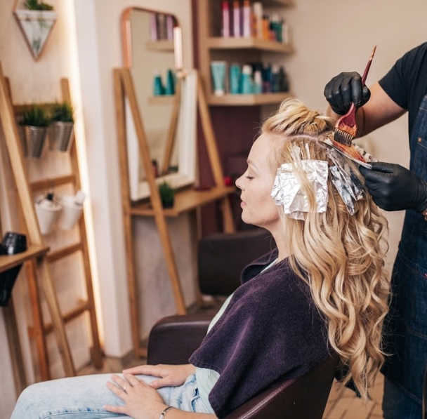 Blonde woman getting foil highlights applied by stylist with brush and bowl in modern hair salon.